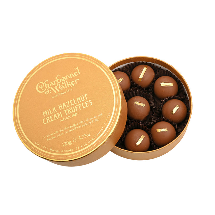 Round gold box with its lid removed, containing seven Hazelnut Cream Milk Chocolate Truffles topped with edible gold leaf.