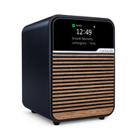 The Ruark R1S Smart Radio is a compact black and wood-finish speaker with a digital display showing the time (12:49) and Spotify info. The front features Ruark branding, emphasizing its Wi-Fi smart radio capabilities.