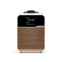 The Ruark R1S Smart Radio by Ruark is a compact, modern tabletop speaker with a wood slat front and white casing. Its digital display shows the time (13:15), now playing "Dakota" by Stereophonics, and Wi-Fi radio for seamless streaming.