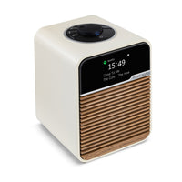 The Ruark R1S Smart Radio features a modern cream body, wood slat front, and black digital display showing 15:49 with Spotify playing Close to Me by The Cure. Enjoy seamless listening with Wi-Fi radio from Ruark.