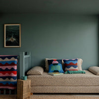 A cozy daybed with patterned pillows and a Donna Wilson Wiggle Square Cushion in red sits by a large window overlooking the beach; a mug and book rest on a small woven table beside it, beneath a framed photo on a muted green wall.