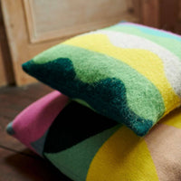 Two Totem Large Square Cushions in green by Donna Wilson—crafted from the softest lambswool and made in the UK, featuring colorful wavy patterns in green, yellow, white, pink, and teal—are stacked on a dark wooden floor next to a light wooden door.