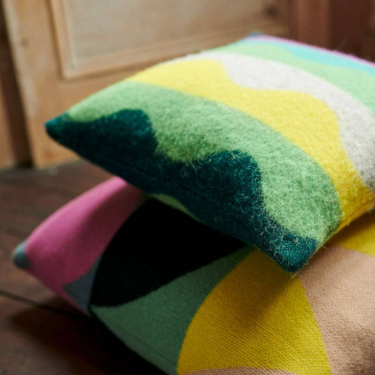 Two vibrant Donna Wilson Wiggle Square Cushions in green—with abstract geometric patterns in shades of green, yellow, white, pink, and teal—rest on a dark wooden floor near a light wooden door; each one is crafted from lambswool in the UK.