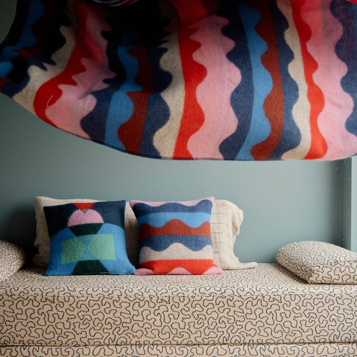A patterned blanket with wavy stripes is being tossed above a sofa adorned with Donna Wilson Wiggle Square Cushions in red, next to a large window overlooking a foggy beach.
