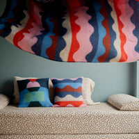 A patterned blanket with wavy stripes is being tossed above a sofa adorned with Donna Wilson Wiggle Square Cushions in red, next to a large window overlooking a foggy beach.