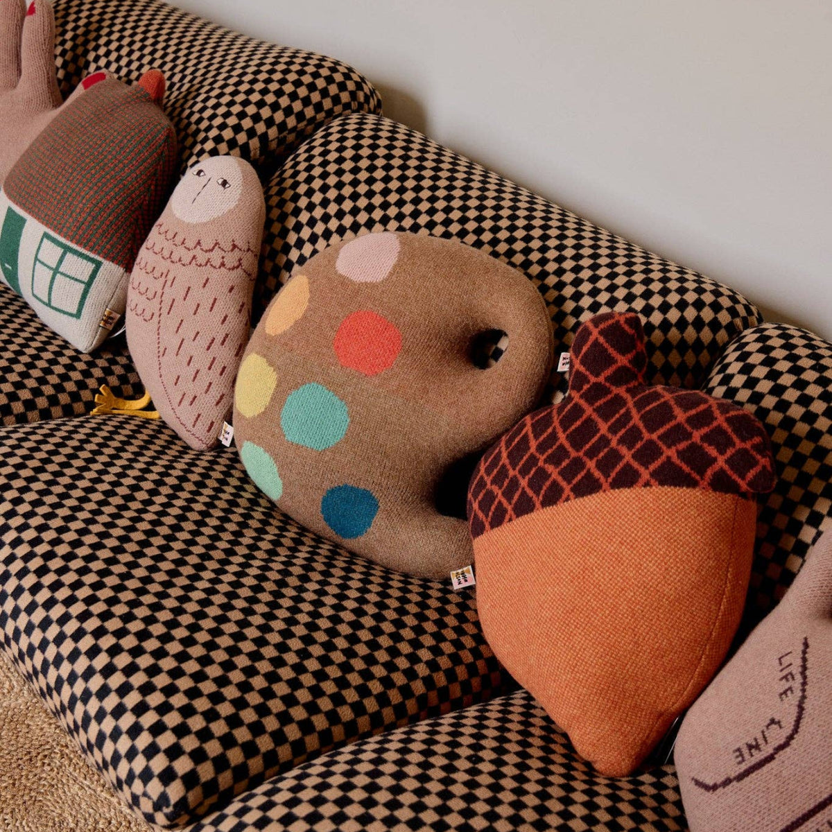 Four decorative cushions, including the Acorn Shaped Cushion by Donna Wilson as well as bear, owl, and polka-dotted rock designs, are arranged on a patterned sofa against a plain wall. These charming lambswool cushions are expertly knitted in Scotland, bringing both comfort and whimsical style to your space.