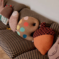 Four decorative cushions, including the Acorn Shaped Cushion by Donna Wilson as well as bear, owl, and polka-dotted rock designs, are arranged on a patterned sofa against a plain wall. These charming lambswool cushions are expertly knitted in Scotland, bringing both comfort and whimsical style to your space.