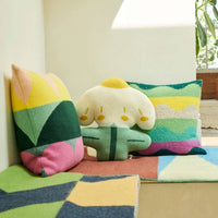 Three colorful, patterned pillows—including the Totem Large Square Cushion in green by Donna Wilson, beautifully crafted in the UK—rest on a bench draped with a geometric-patterned blanket. Sunlight streams through a window, illuminating the cozy, modern room adorned with potted plants in the background.