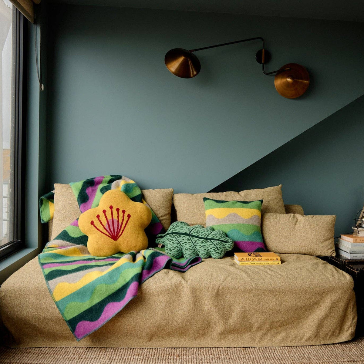 Cozy room with a beige sofa, colorful pillows, and a Donna Wilson Wiggle Square Cushion in green, made in the UK. Brass wall lights, stacked books, and art prints decorate the blue-green walls. A large window lets in natural light.
