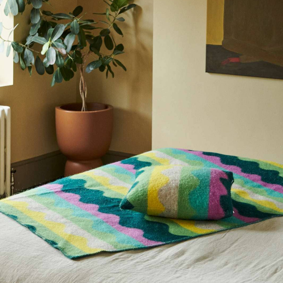A cozy bedroom with a potted plant, beige walls, abstract wall art, and a bed covered with a colorful, wavy-patterned blanket and the Donna Wilson Wiggle Square Cushion in green—each piece thoughtfully made in the UK.