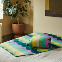 A cozy bedroom with a potted plant, beige walls, abstract wall art, and a bed covered with a colorful, wavy-patterned blanket and the Donna Wilson Wiggle Square Cushion in green—each piece thoughtfully made in the UK.