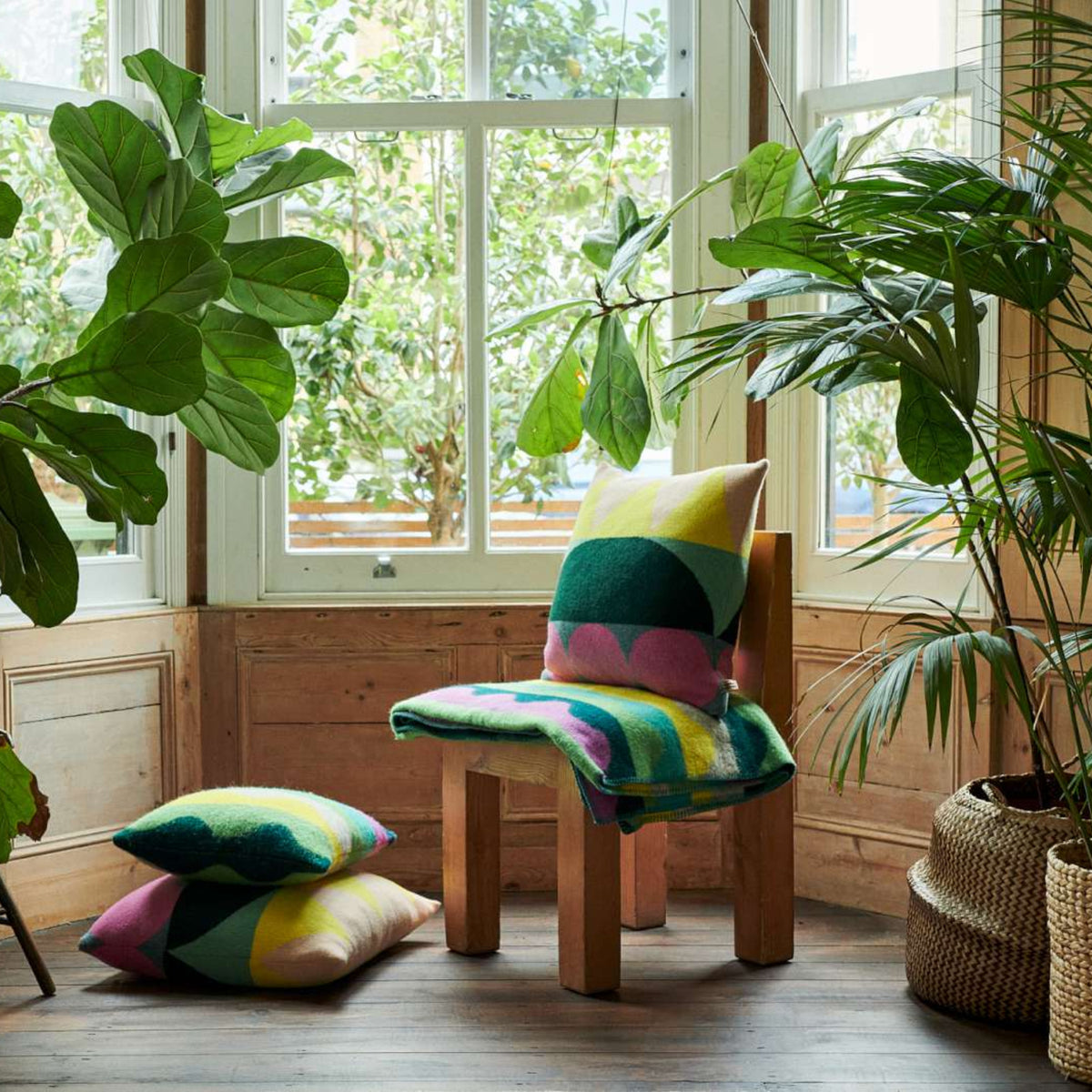 A cozy sunlit room with large windows, leafy green plants, a wooden chair adorned with colorful pillows and a soft lambswool cushion, a blanket draped over the side, a pillow on the floor, a small stool with a plush toy, and a woven basket. The inviting space features thoughtful touches like the Totem Large Square Cushion in Green by Donna Wilson—crafted in the UK—which adds both comfort and charm to this peaceful retreat.