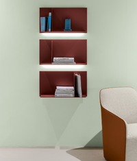 Three red Avenue Library Shelving units by Kristalia, each H32cm, displaying books and a blue object, are mounted on a light green wall above a beige and brown armchair.