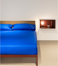 A neatly made bed with bright blue sheets and pillows is next to a wall niche featuring a lit bulb lamp and a glass jar, complemented by the Kristalia Avenue Library Shelving, H32cm, which enhances the calm, modern feel of this room with white walls and a beige floor.
