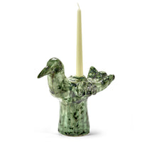 The Cosas Lindas Candle Holder by Serax is a green ceramic piece shaped like a bird, adorned with delicate carved details. Designed to hold a tall, pale yellow taper candle on its back, it makes a charming addition to any nature-inspired decor.