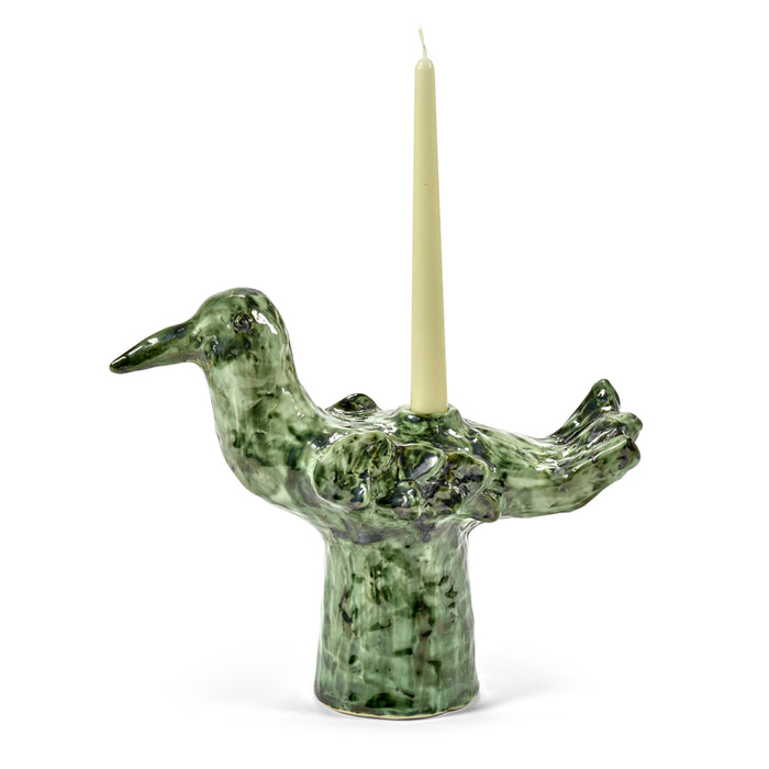 The Cosas Lindas Candle Holder by Serax is a green ceramic piece shaped like a bird, with intricately carved details and a mottled texture on its body and head for added nature-inspired charm. A cream-colored tapered candle is elegantly inserted vertically in the center of the bird’s back.