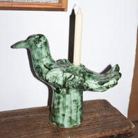 The Cosas Lindas Candle Holder by Serax is a nature-inspired decor piece, crafted from green ceramic and shaped like a bird with delicate carved details. Designed to hold a tall white candle on its back, it sits gracefully on a wooden surface against a white wall.