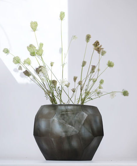 The Cubistic Vase Indigo-Smoke Grey by Guaxs, a geometric mouth-blown glass vase handmade in Germany, elegantly displays long-stemmed wildflowers with white and green blooms against a softly lit white background.