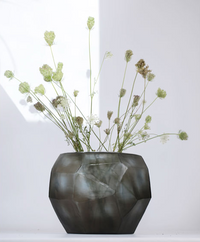 The Cubistic Vase Indigo-Smoke Grey by Guaxs, a geometric mouth-blown glass vase handmade in Germany, elegantly displays long-stemmed wildflowers with white and green blooms against a softly lit white background.