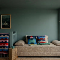 Two Donna Wilson Shape Shift cushions with abstract, colorful geometric and wavy patterns resting on a patterned daybed.
