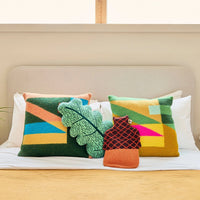 Two Tilt Cushion, Green square geometric cushions, a leaf-shaped pillow, and a small acorn-shaped hot water bottle cover arranged on a bed, all crafted from pure new wool.