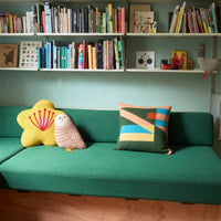 A star-shaped pillow, an owl-shaped pillow, and a rectangular Tilt Cushion, Peach, on a green sofa below two shelves of books.