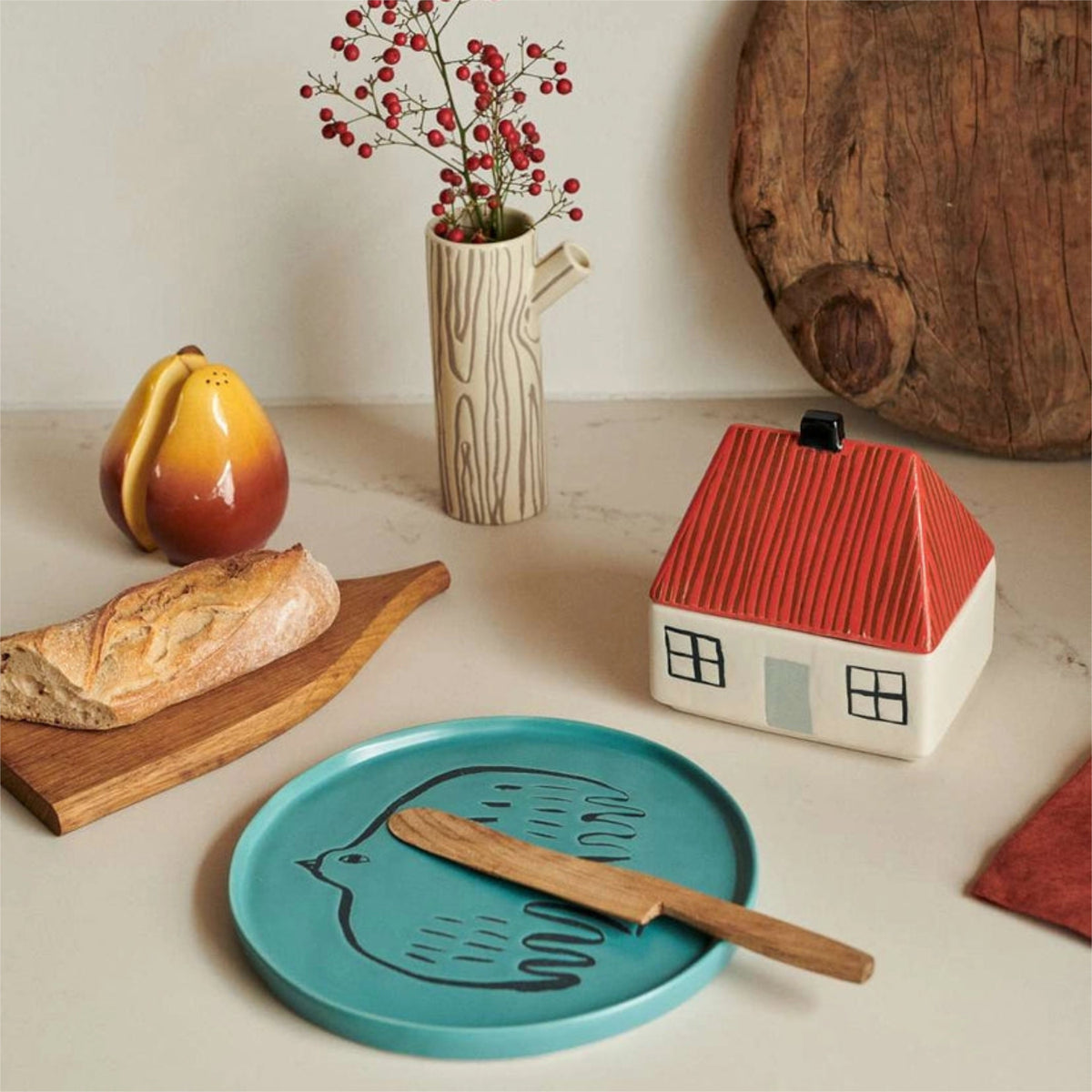 A table with a loaf of bread, pear-shaped salt and pepper shakers, a vase filled with red berries, a plate featuring a bird design, a wooden butter knife, and the charming Donna Wilson House Butter Dish by Donna Wilson.