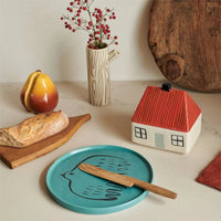 A table with a loaf of bread, pear-shaped salt and pepper shakers, a vase filled with red berries, a plate featuring a bird design, a wooden butter knife, and the charming Donna Wilson House Butter Dish by Donna Wilson.