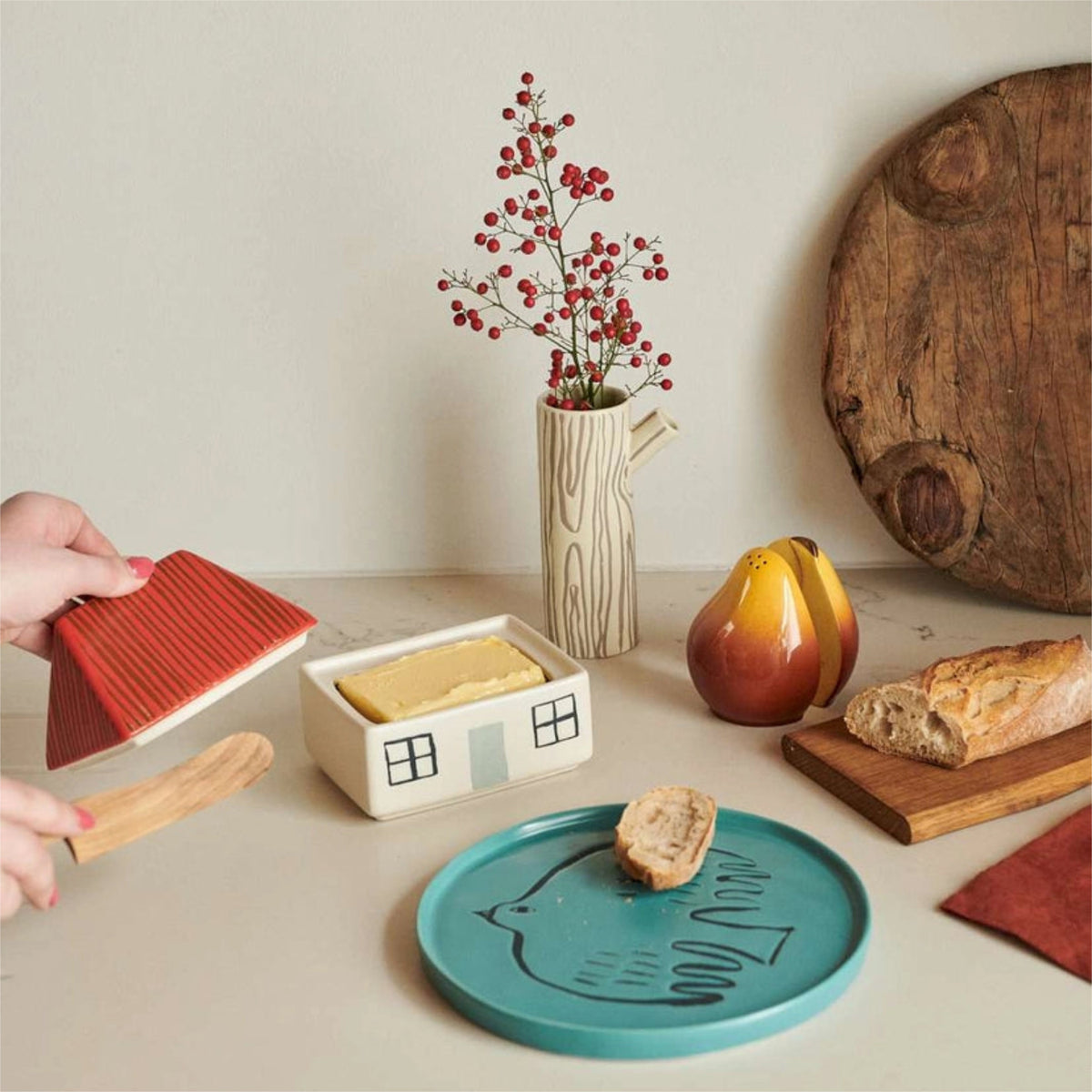 A hand opens the Donna Wilson House Butter Dish, a delightful stoneware piece by Donna Wilson shaped like a cozy house. Nearby rest a bread-filled plate, a vase with red berries, a wooden board holding a baguette, and a pear-shaped object on a light tabletop.