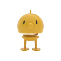 Yellow round Hoptimist Bumble desk toy with a spring-mounted head, two simple eyes, a round nose, and two feet.