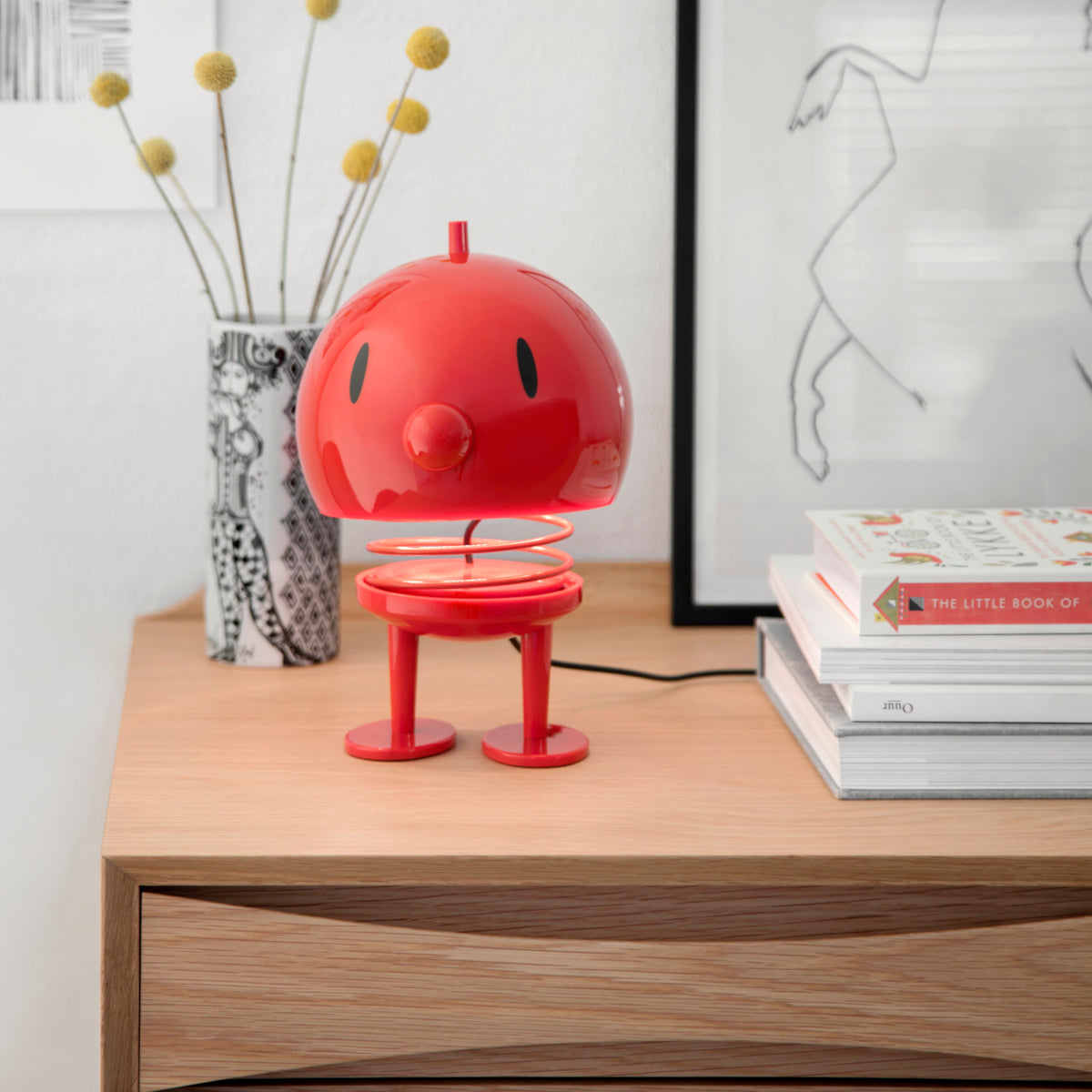 A small, red, spring-mounted Hoptimist Bumble Desk Toy with a round head and feet, standing on a wooden desk as a piece of Danish design.