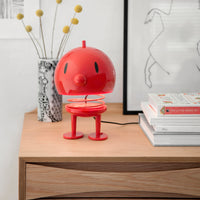 A small, red, spring-mounted Hoptimist Bumble Desk Toy with a round head and feet, standing on a wooden desk as a piece of Danish design.