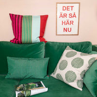 A green couch with three colorful pillows, including the Josefina Cushion by World Of Craft—handwoven by Guatemalan artisans—sits beneath a framed sign reading DET ÄR SÅ HÄR NU. An open magazine lies on the couch, and the background wall is a soft pastel shade.