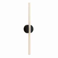 The Kilter Wall Light 640mm by Tala is a modern wall sconce featuring a long, vertical, cylindrical white borosilicate glass fixture elegantly mounted on a round black base against a plain white background—ideal for sophisticated vanity lighting.
