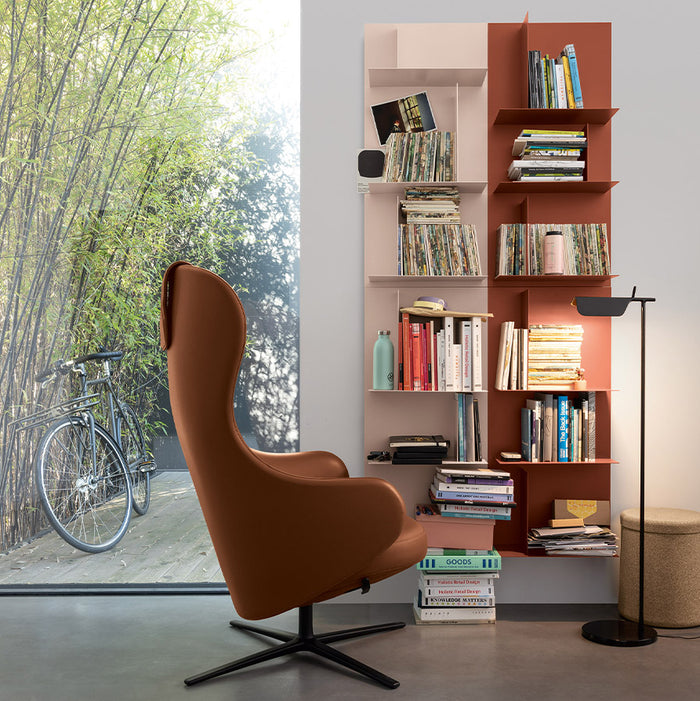A modern reading nook featuring Kristalia’s Avenue Library Shelving H96cm used as a contemporary bookcase filled with books and records, complemented by a brown swivel chair, a floor lamp, and a large window overlooking a bicycle and bamboo outside.
