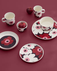 An assortment of plates, bowls, cups—including the Marimekko Oiva / Unikko Mug 4dl—paired with a small dish, all featuring bold red, black, and white Unikko floral patterns. The tableware is set on a coordinated red surface with two red apples, creating a striking and cohesive display.