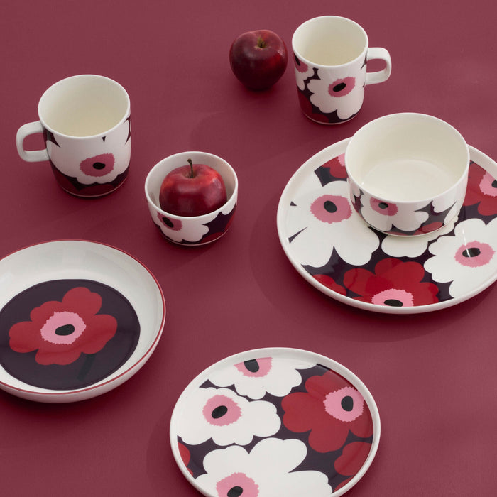 An assortment of plates, bowls, cups—including the Marimekko Oiva / Unikko Mug 4dl—paired with a small dish, all featuring bold red, black, and white Unikko floral patterns. The tableware is set on a coordinated red surface with two red apples, creating a striking and cohesive display.