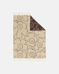 The Marimekko Piirto Unikko Blanket is a beige wool throw featuring a large, bold floral pattern outlined in brown. One corner is folded to reveal the reverse side with the same design in inverted colors. Inspired by Marimekko’s classic Unikko motif, this blanket is finished with fringe on the edges and is crafted from responsibly sourced fibers that meet the Responsible Wool Standard.
