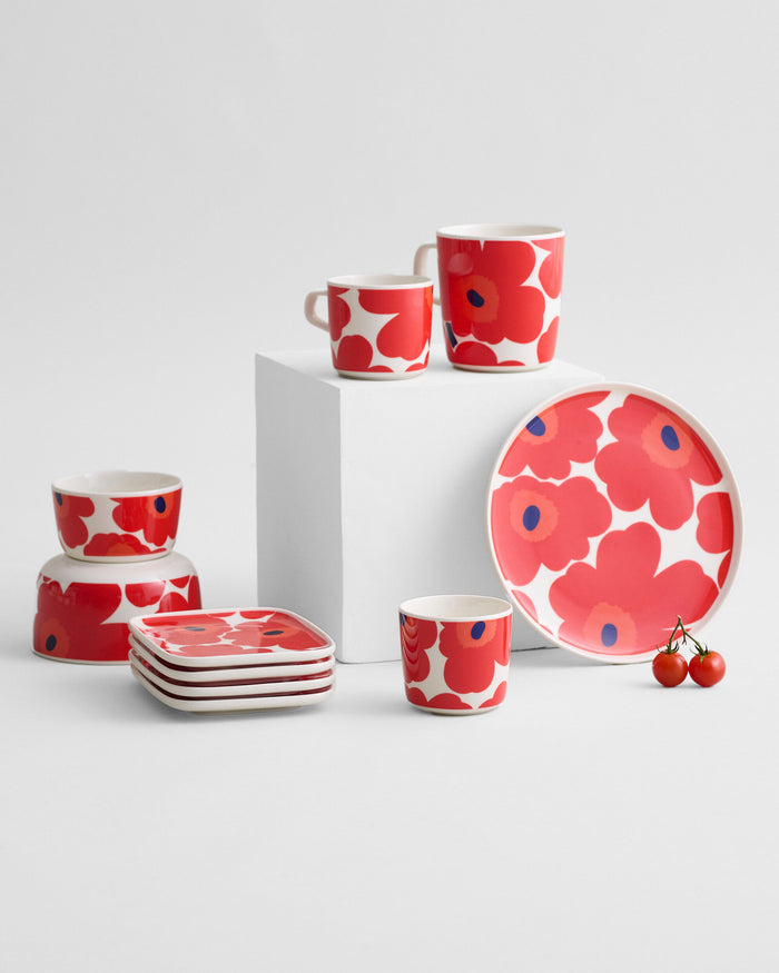 A set of red and white dishware featuring bold red floral patterns—including plates, mugs, and the Marimekko Oiva Unikko Bowl (50cl) in red—arranged on a white surface with two cherry tomatoes nearby.