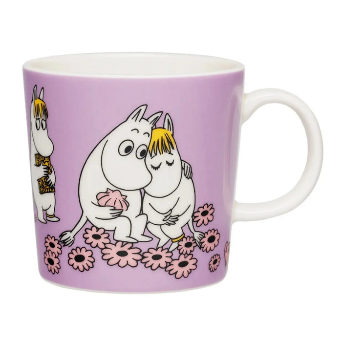 The Moomin Mug - Sweethearts by Arabia is a lavender ceramic mug from the Moomin collection, inspired by Tove Jansson, and depicts cartoon hippopotamus-like characters—one embracing another and a third standing nearby—sitting among pink flowers.