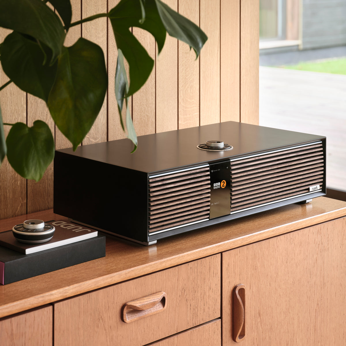 The Ruark R410 All-In-One Music System by Ruark, featuring a sleek black and wood design, sits on a wooden cabinet in a stylish room with wood-paneled walls, large windows, and a hanging plant on the left.