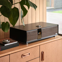 The Ruark R410 All-In-One Music System by Ruark, featuring a sleek black and wood design, sits on a wooden cabinet in a stylish room with wood-paneled walls, large windows, and a hanging plant on the left.