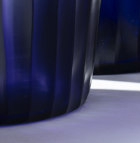 Close-up of the Ligne Roset Purple Rain Vase, a dark blue translucent decorative piece with vertical ridges, displayed on a light surface. Bright lighting casts striking contrasts in the background.