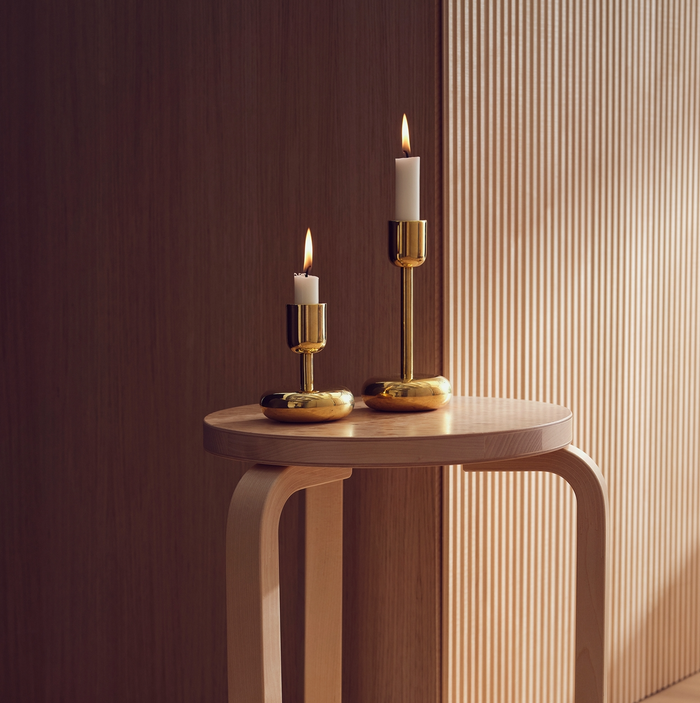 Two white candles in Nappula Brass Candle Holders (2 Pack) burn on a light wooden round table, with vertical wooden paneling and soft, warm lighting in the background.