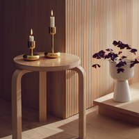 A round wooden stool holds two lit candles in Nappula Brass Candle Holders, 2 Pack. Next to it, a white vase with dark purple leaves sits on a wooden step beside a wall with vertical wood paneling, all softly illuminated.