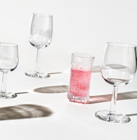 Four Ultima Thule Sparkling Wine Glasses are arranged on a white surface: three are empty, highlighting Nordic design, while one tumbler holds pink liquid. Distinct shadows appear beneath each glass in the bright light.