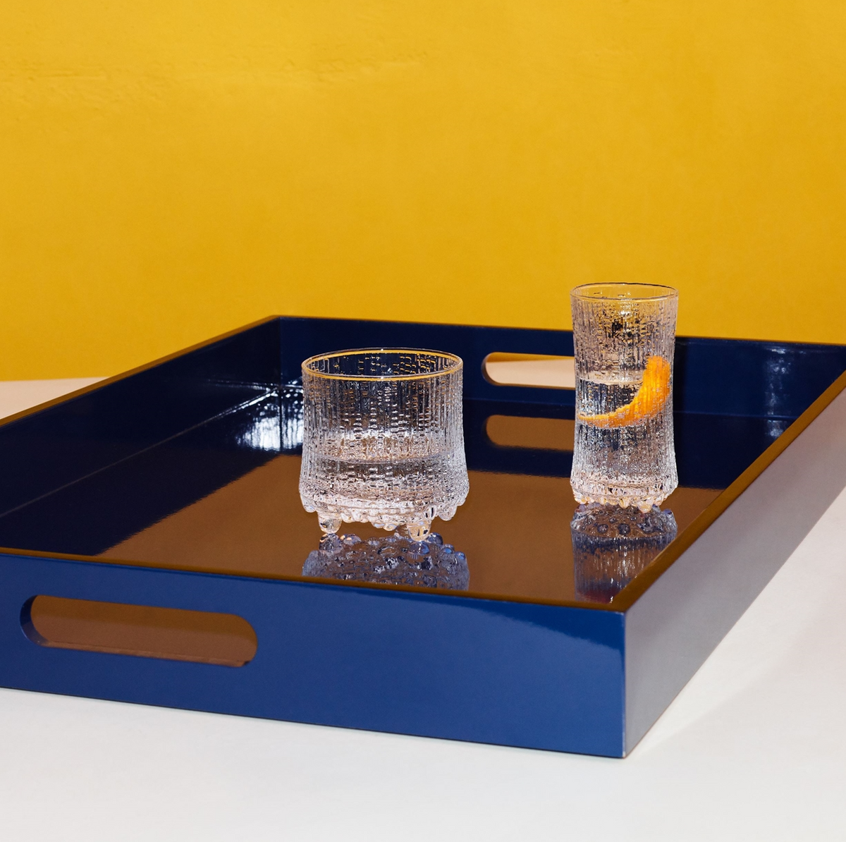 A glossy dark blue tray holds two textured clear Ultima Thule Sparkling Wine Glasses—one short and empty, one tall with water and an orange peel—highlighting Nordic design against a yellow background.
