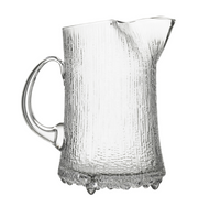 The Ultima Thule Pitcher 150cl features mouth-blown glass with a textured surface, curved handle, and wavy spout. Its base is decorated with small raised glass beads, reflecting Iittala’s signature style.