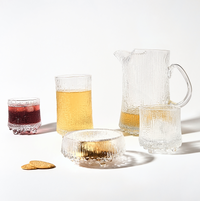 A set of textured Iittala glassware, featuring the Ultima Thule Pitcher 150cl, two glasses with yellow drinks, a glass with a red drink, a low bowl with a yellow drink, and two round crackers on a white surface.
