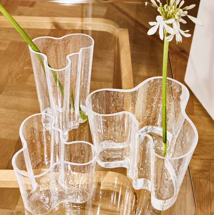 Three clear Alvar Aalto Bubble Vases with organic, wavy shapes are partially filled with water, each showcasing a single long-stemmed white flower. The vases sit on a wooden tray placed on a wooden surface.
